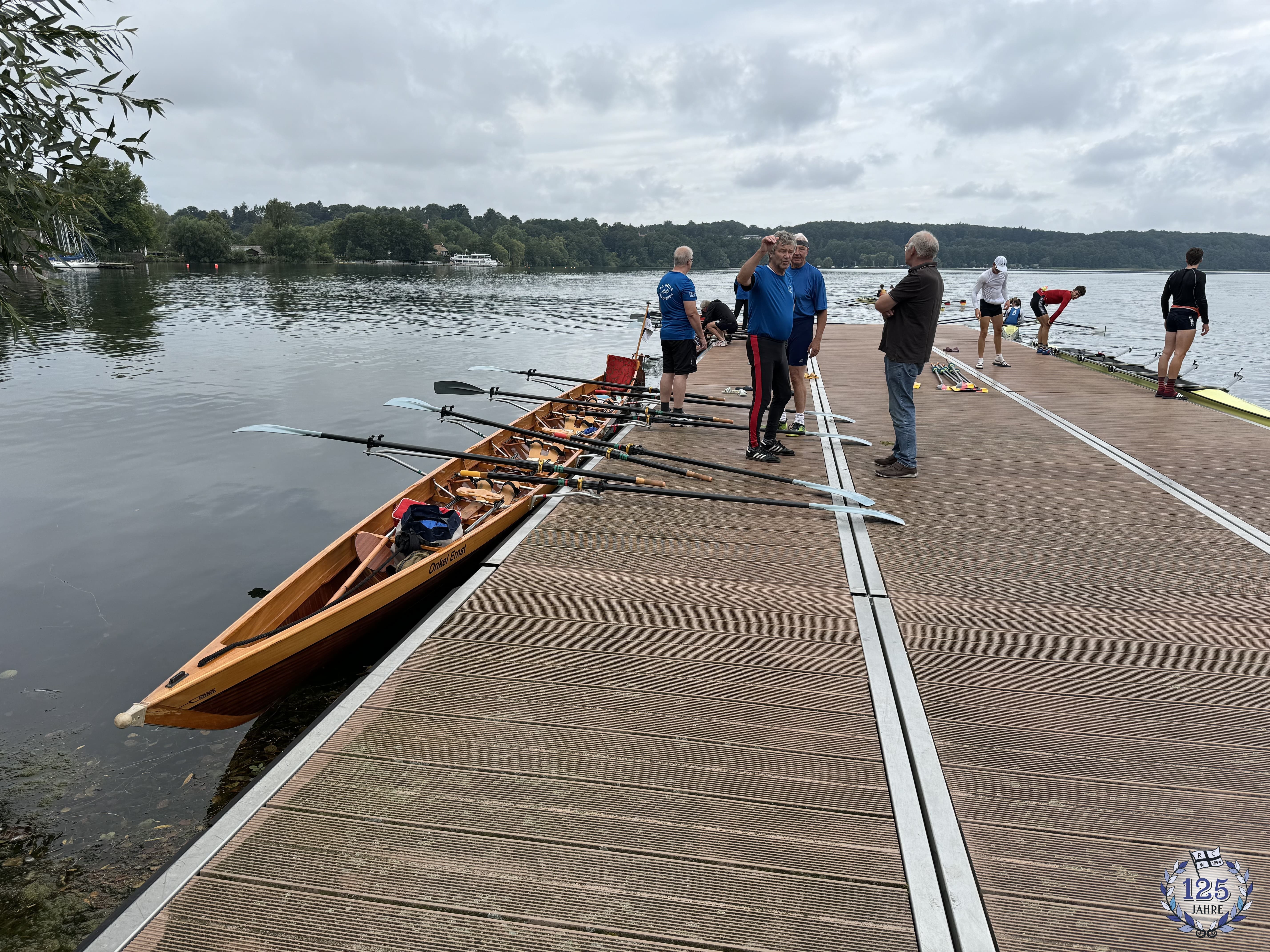 Links ein Gig-Vierer am Steg bereit zur Abfahrt. Auf dem Steg stehen einige Ruderer. Rechts vom Steg bereiten Ruderer ihr Rennboot vor.