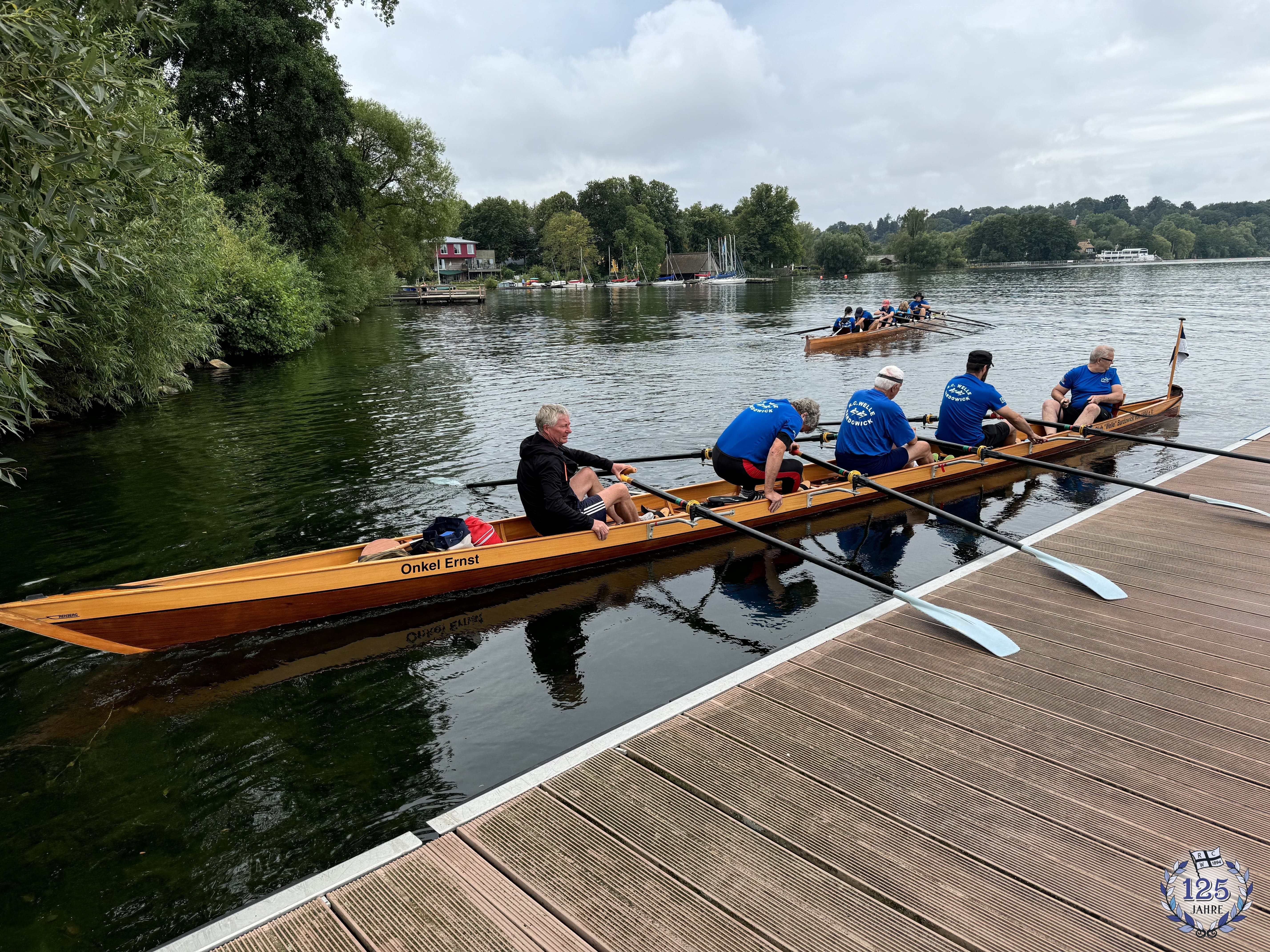 Ein Team von 5 Ruderern in einem Holzboot bereitet sich darauf vor, auf einem See zu rudern. Das Boot liegt neben einem Steg, während im Hintergrund ein weiteres Team rudert. Bäume und Häuser säumen das Seeufer.