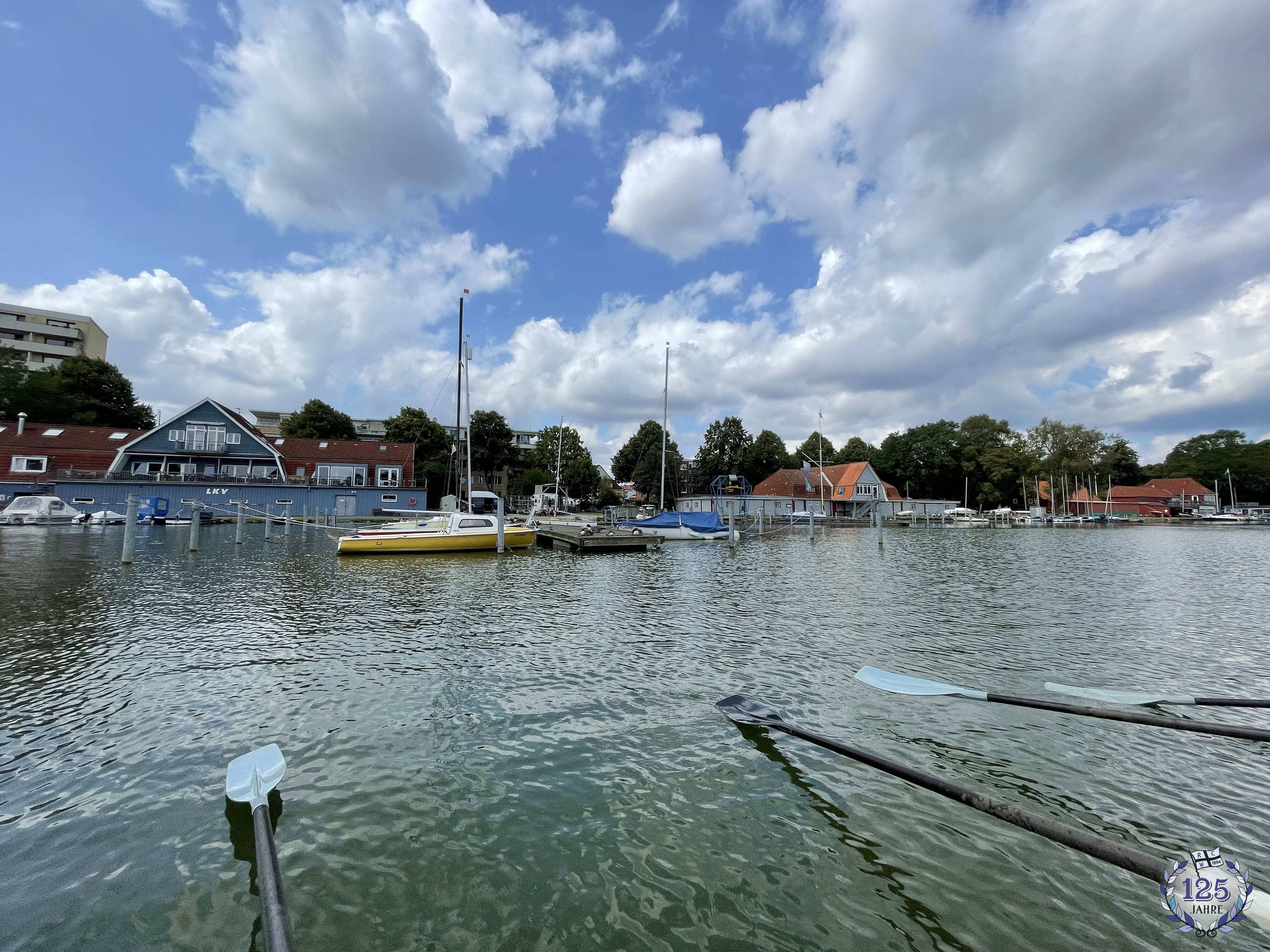 Ein Bootsanleger mit mehreren Segelbooten und kleineren Wasserfahrzeugen liegt ruhig im Wasser. Im Vordergrund ragen zwei Ruderblätter ins Bild, während im Hintergrund Häuser mit roten und blauen Dächern zu sehen sind. Der Himmel ist teils bewölkt mit blauen Abschnitten.