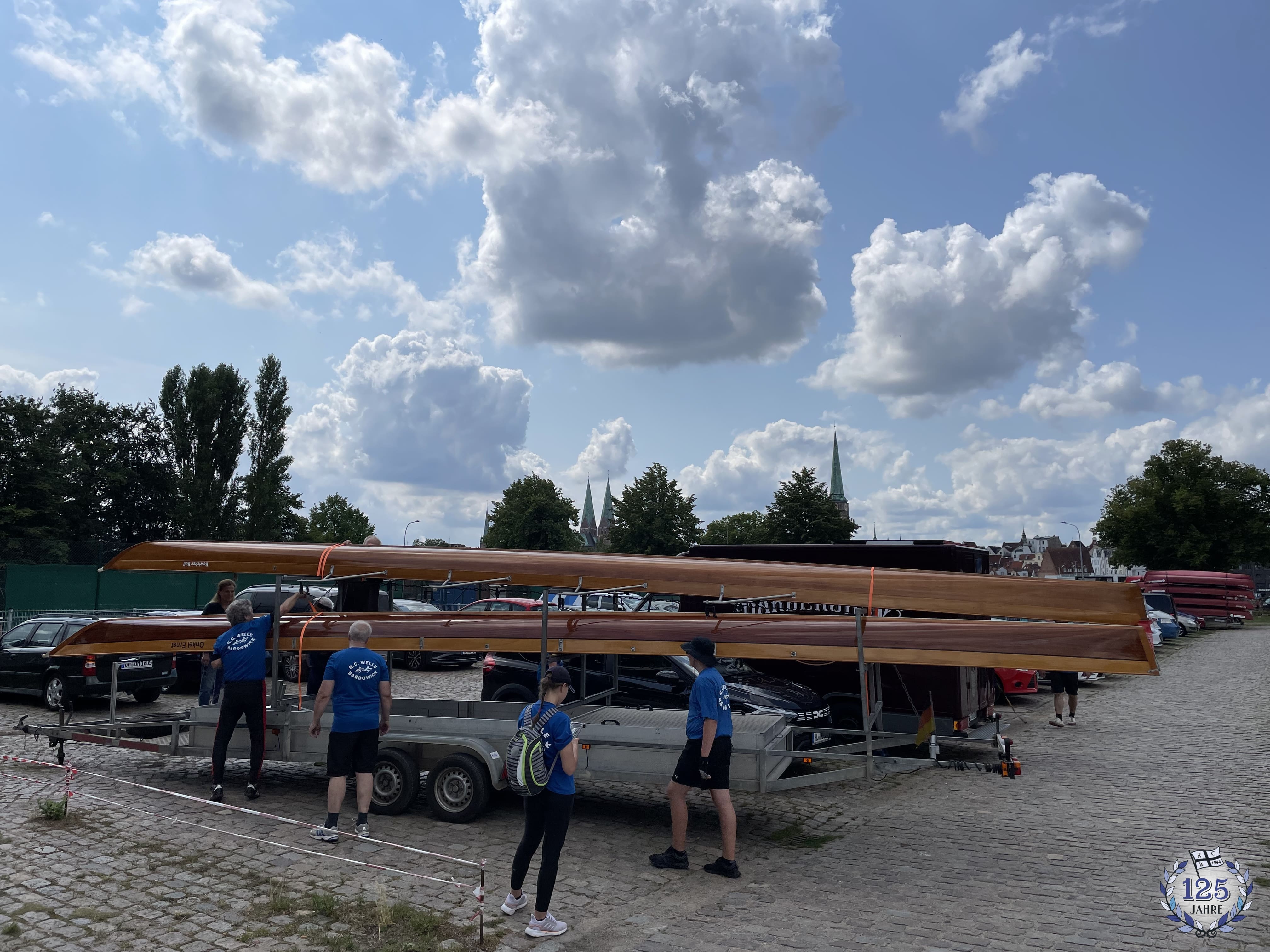 Mehrere Personen in blauen T-Shirts laden lange, hölzerne Ruderboote auf einen Anhänger. Sie stehen auf einem gepflasterten Platz, im Hintergrund sind Bäume und Kirchturmspitzen vor einem teilweise bewölktem Himmel zu sehen.