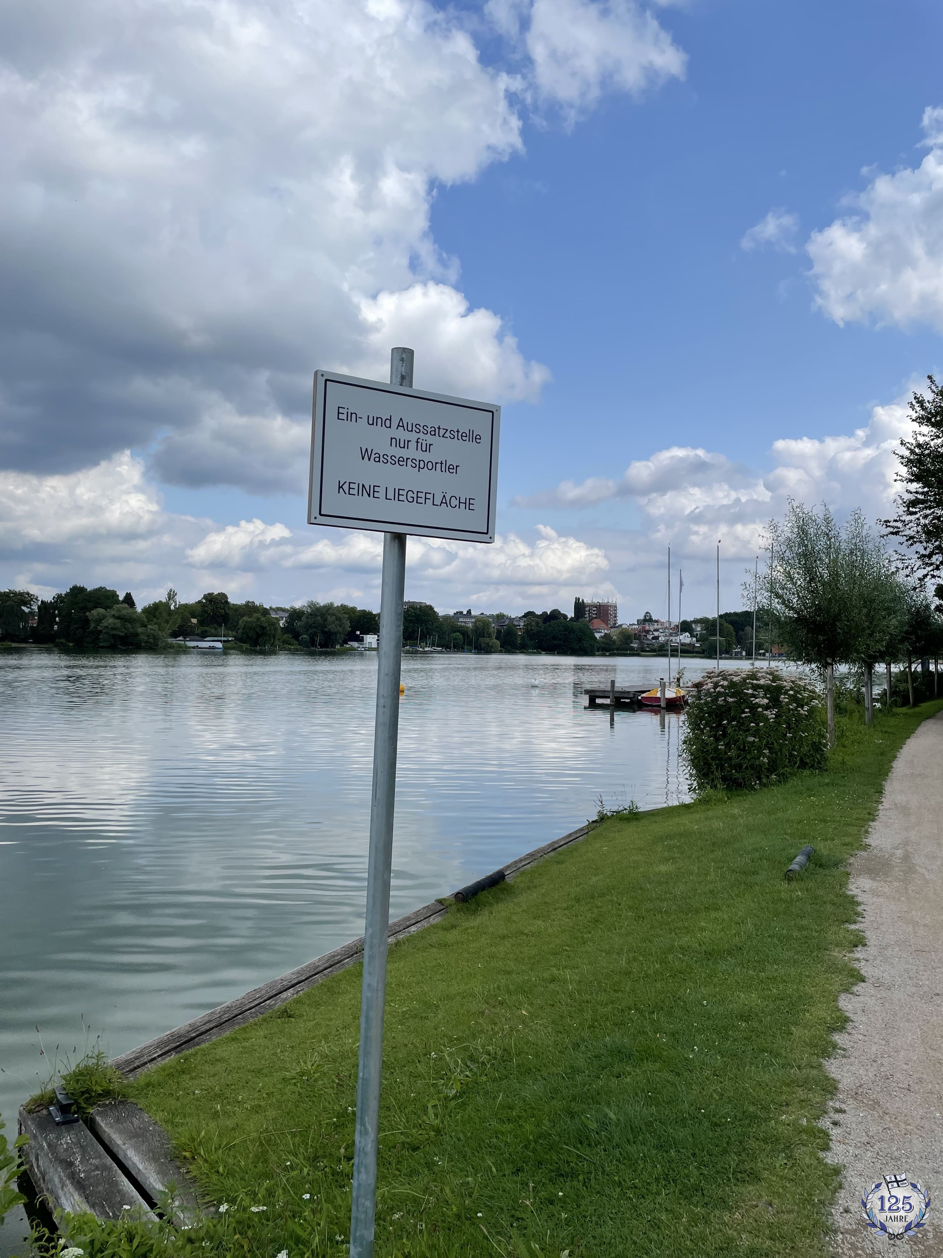 Im Bild ist ein ruhigen Gewässer zu sehen. Im Vordergrund steht ein weißes Schild mit der Aufschrift 'Ein- und Aussatzstelle nur für Wassersportler - KEINE LIEGEFLÄCHE'. Im Hintergrund sind einige Häuser zwischen grünen Laubbäumen zu sehen.