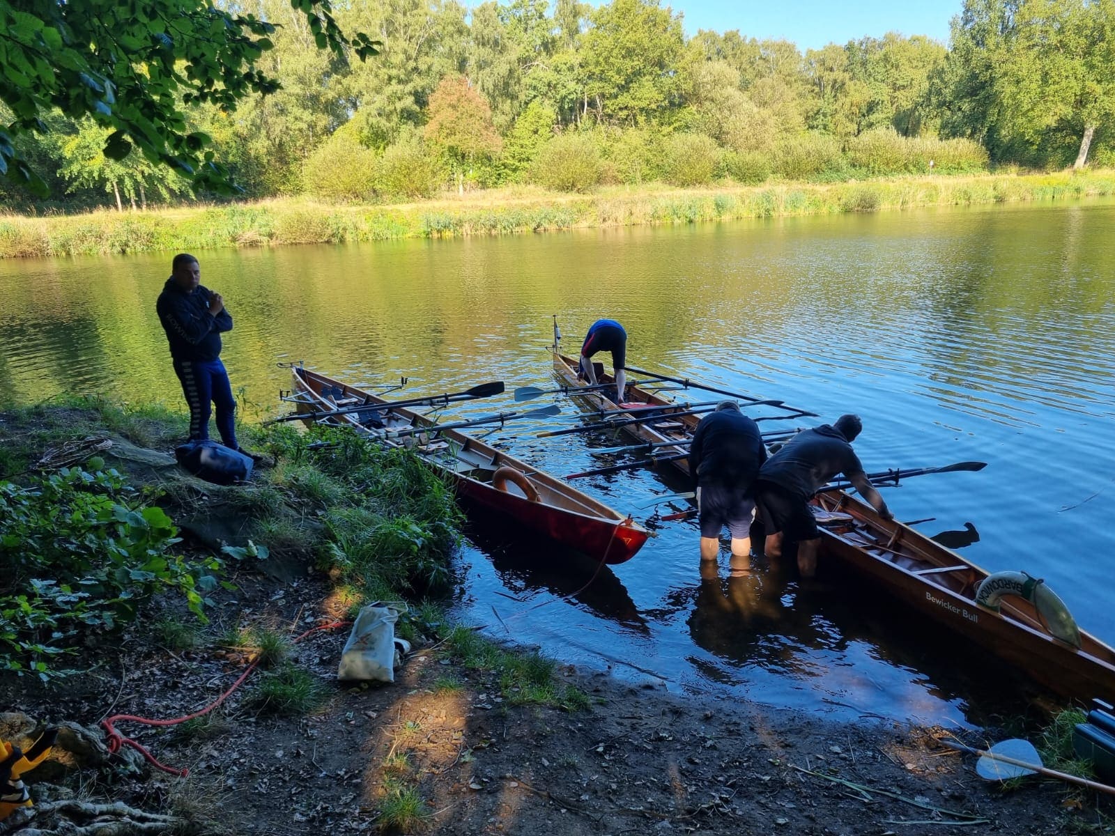 Eine Gruppe von Ruderern am Ufer des Elbe-Lübeck-Kanals. Zwei Boote liegen im Wasser, während einige Personen diese zur Fahrt vorbereiten. Die Umgebung ist naturbelassen. Ein Mann steht am Ufer und beobachtet die Szene.