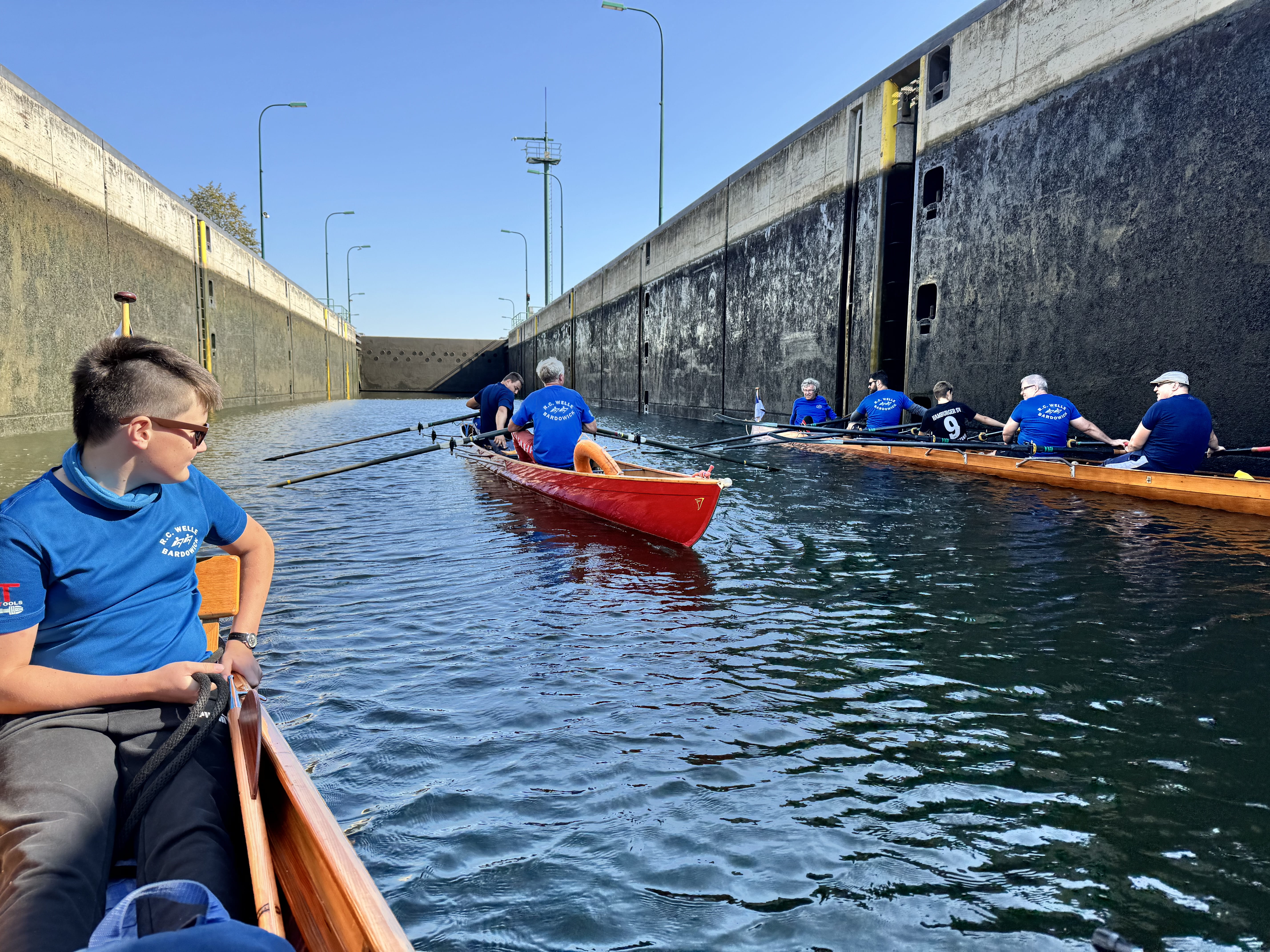 Drei  besetzte Ruderboote inmitten einer großen Schleuse. Am Bildrand sind die feuchten Schleusenwände zu sehen. Im Hintergrund ein Schleusentor.