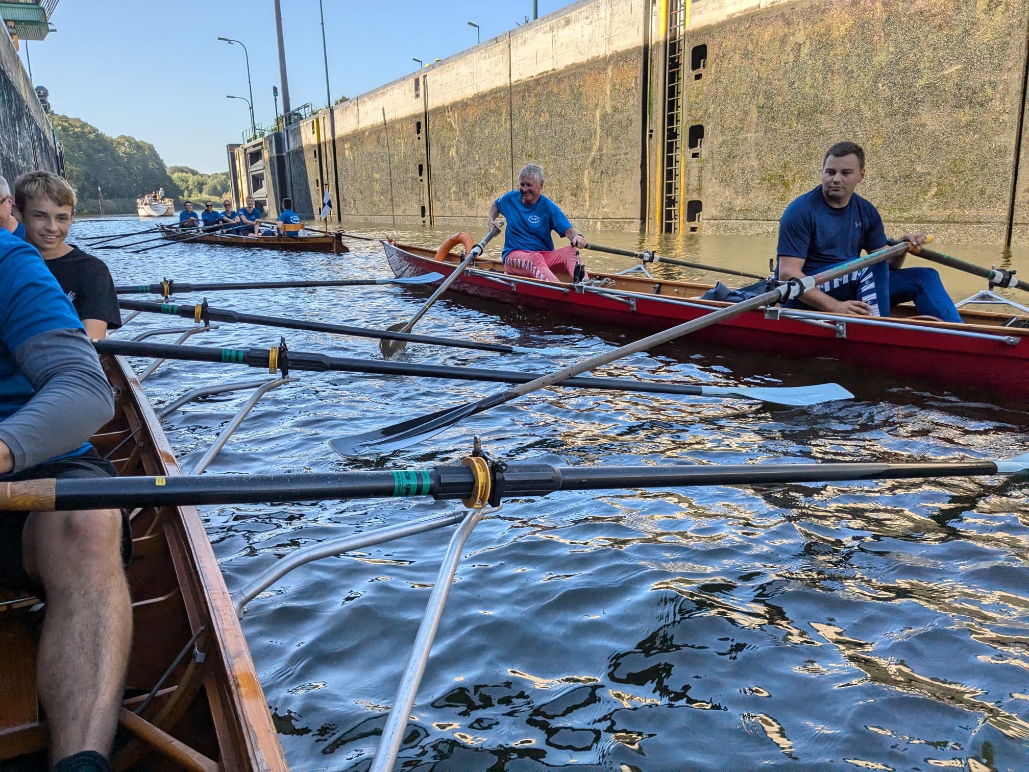 Eine Gruppe von Ruderern sitzt in mehreren Ruderbooten, die in einer Schleuse auf dem Wasser treiben. Die Skulls sind eingetaucht. Im Hintergrund ist die Wand der Schleuse sichtbar, während die Sonne auf das Wasser scheint.