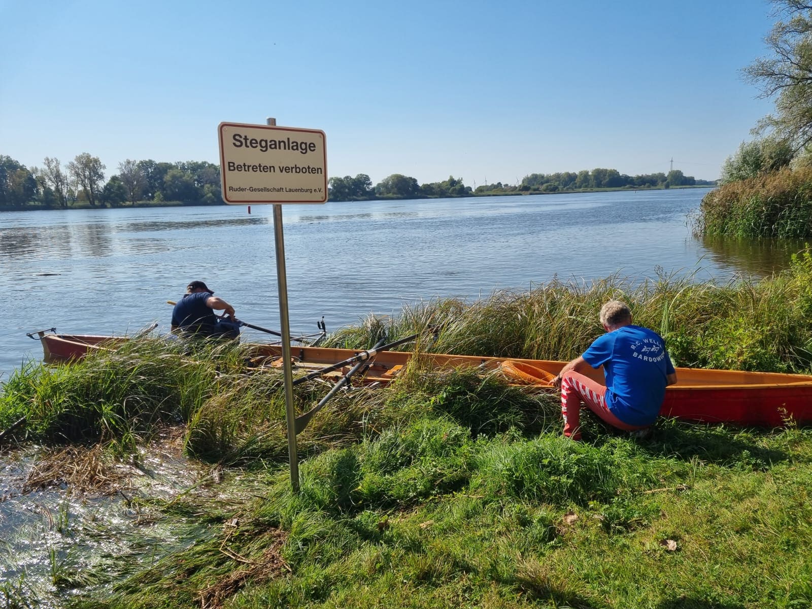 Zwei Personen hocken an einem Ruderboot, das am Elbufer in einer Buhne festgemacht ist. Ein Schild am Ufer weist auf ein Betretungsverbot der Steganlage hin. Die Umgebung ist ruhig, und das Wasser des Flusses spiegelt den blauen Himmel wider.