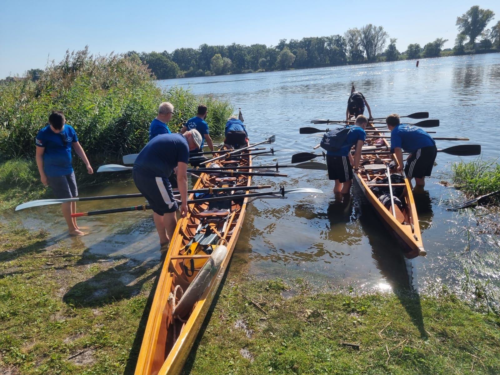 Eine Gruppe von Ruderern steht barfuß im flachen Wasser und bereitet Ihre Boote vor. Die Boote liegen am Elbufer in einer Buhne.